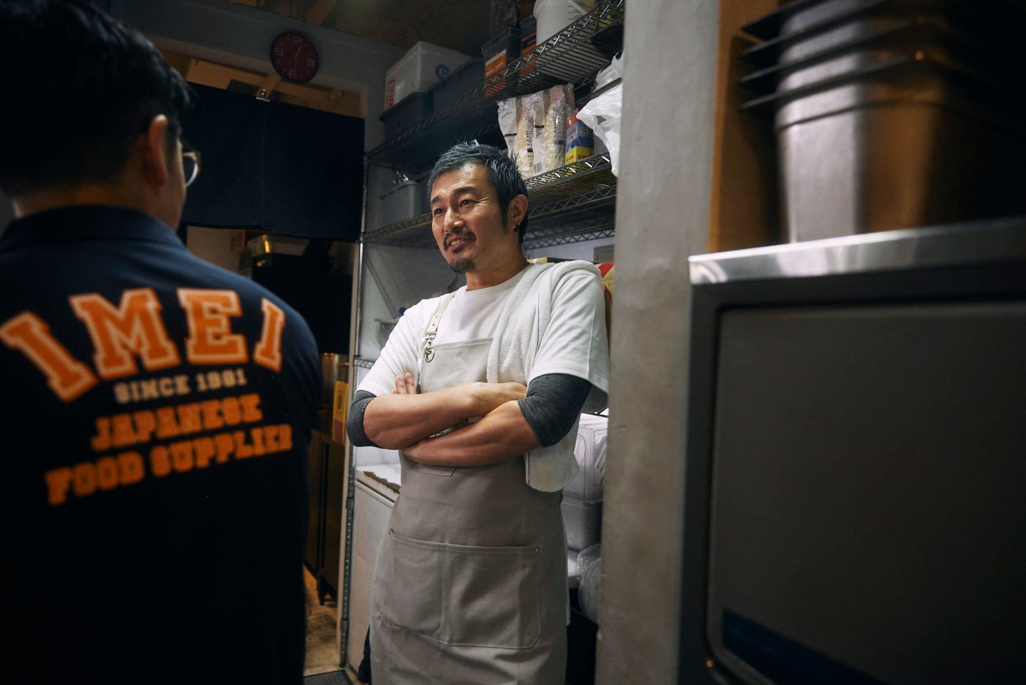 A man in a white apron stands with his arms crossed in a small kitchen, surrounded by shelves and appliances. He has short, dark hair and a mustache, and wears a white apron over.