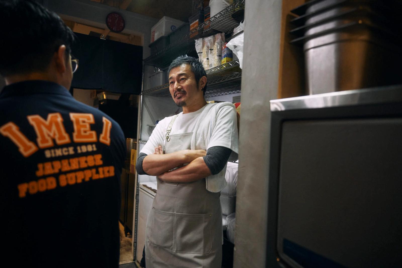 A man in a white apron stands with his arms crossed in a small kitchen, surrounded by shelves and appliances. He has short, dark hair and a mustache, and wears a white apron over.