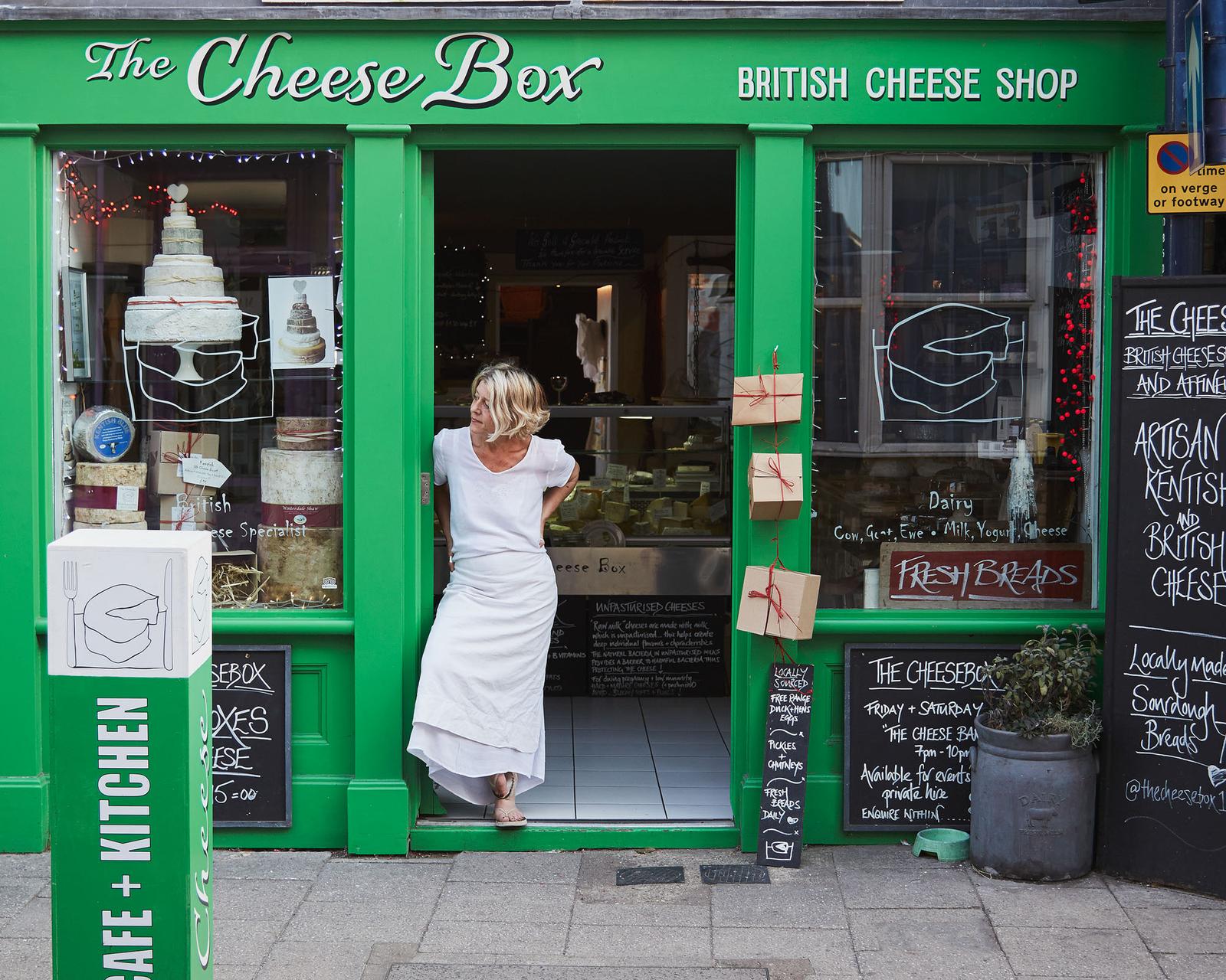A woman in a white dress standing in the doorway of a green-painted shop with a sign reading 'The Cheese Box' and a window display of various types of cheese and a wedding cake.