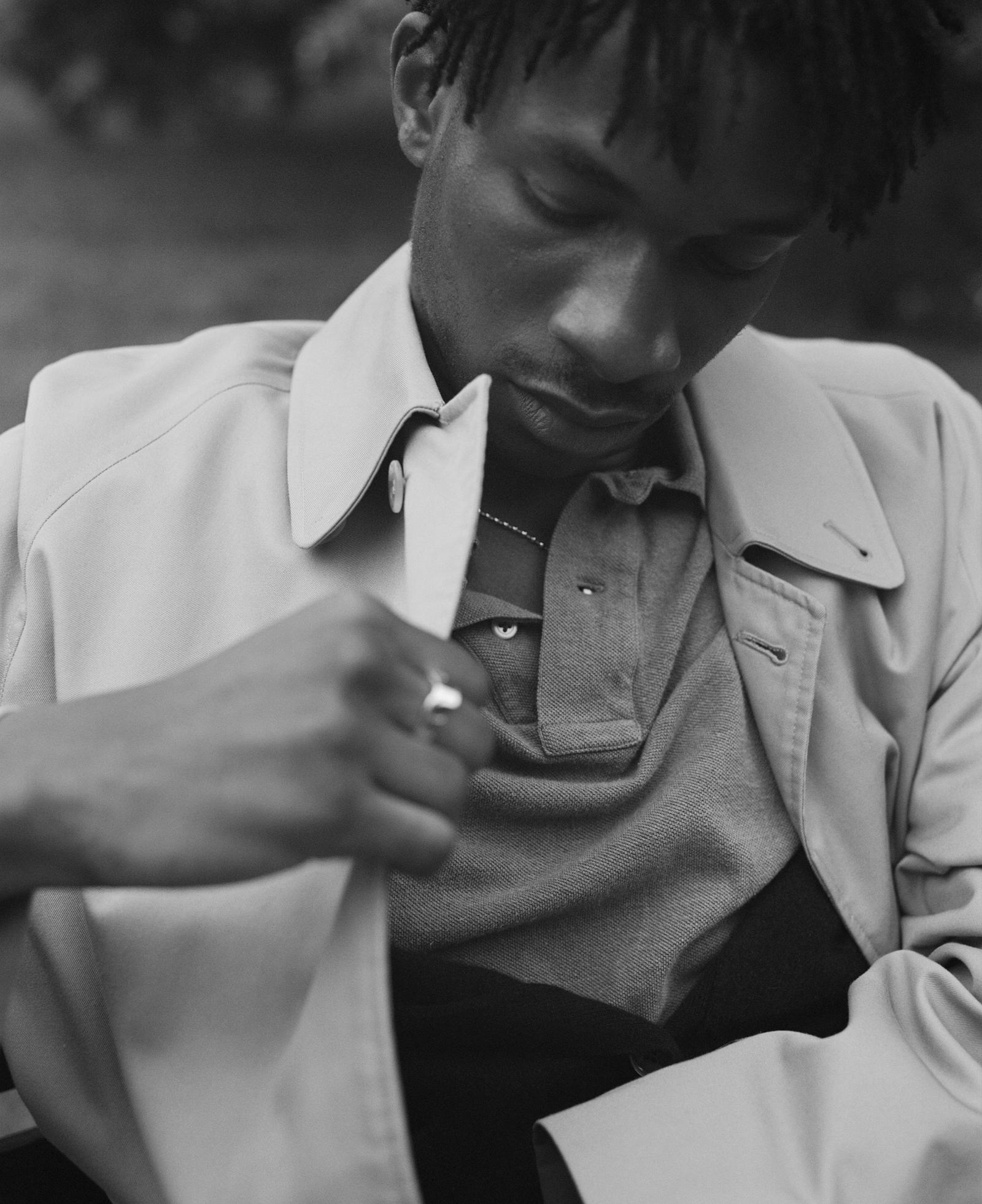 A man in a trench coat and polo shirt, possibly adjusting his shirt buttons.
