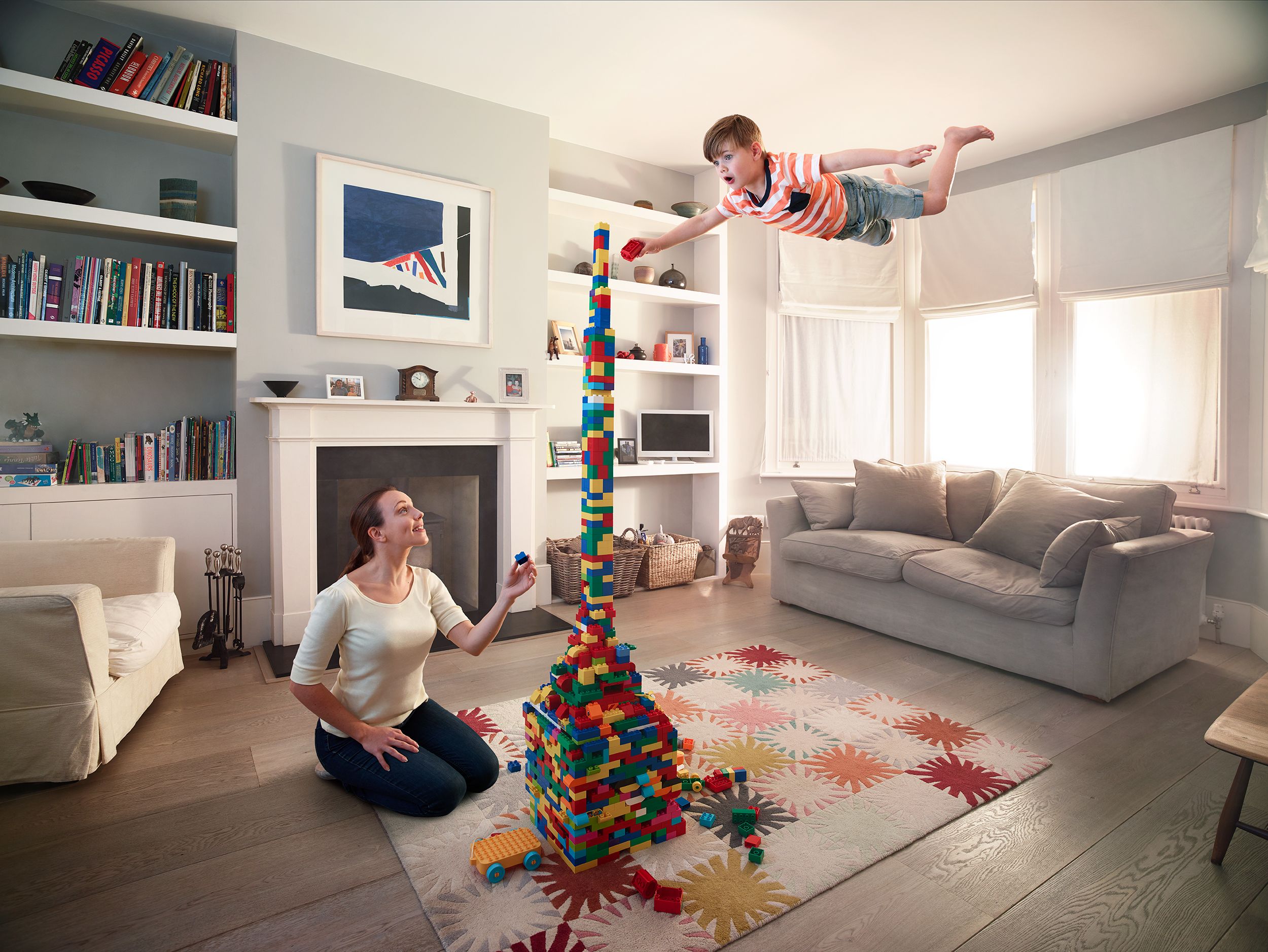 A woman and a young boy are playing with a large tower of colorful LEGO bricks in a living room.