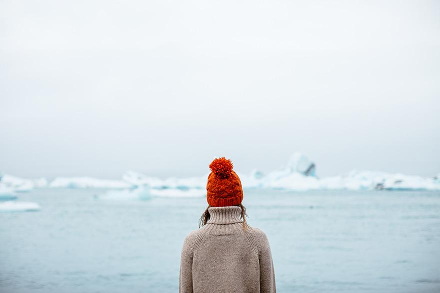 A woman in a red hat and grey coat stands at the edge of a frozen lake, looking out at a vast expanse of ice and snow.