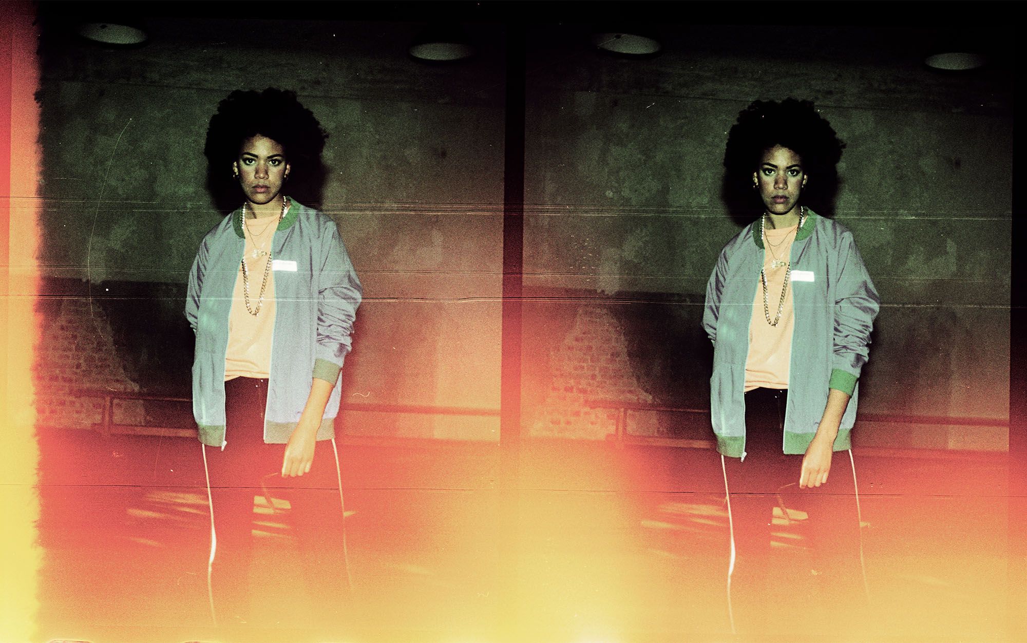 A young woman with curly hair and a serious expression, wearing a light blue jacket, a white t-shirt, and black pants, standing in front of a dark background.