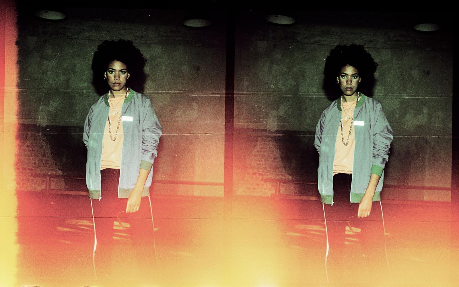 A young woman with curly hair and a serious expression, wearing a light blue jacket, a white t-shirt, and black pants, standing in front of a dark background.