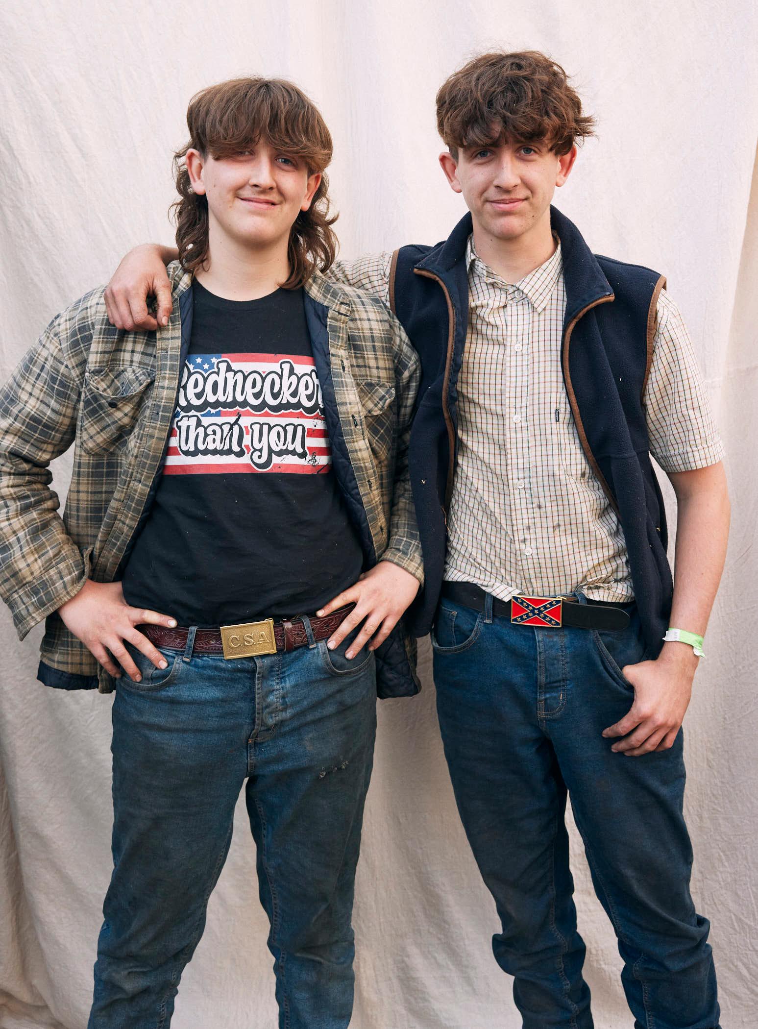 Two young men, likely brothers, stand together with their arms around each other, smiling and looking directly at the camera. They have shaggy, curly hair and are dressed in.