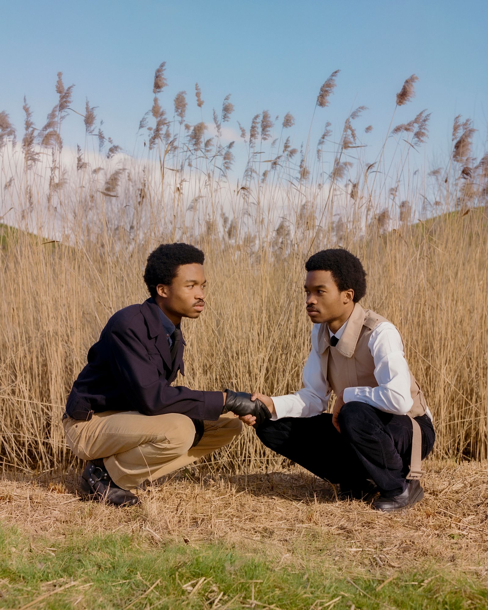 Two men sit in a field, holding hands and looking at each other with serious expressions.