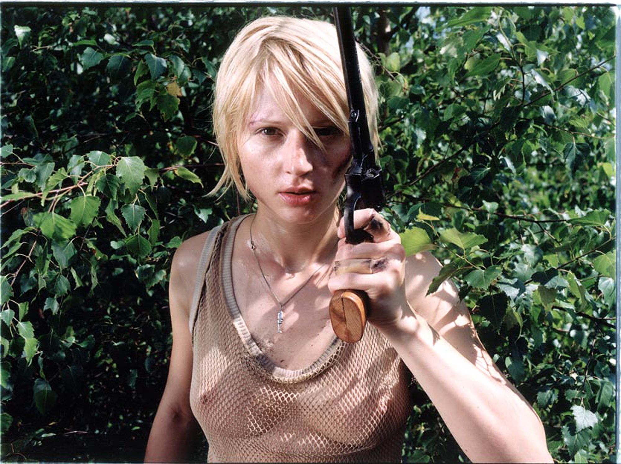 A woman with a short, blonde haircut and a tank top, holding a gun in a forest-like setting.