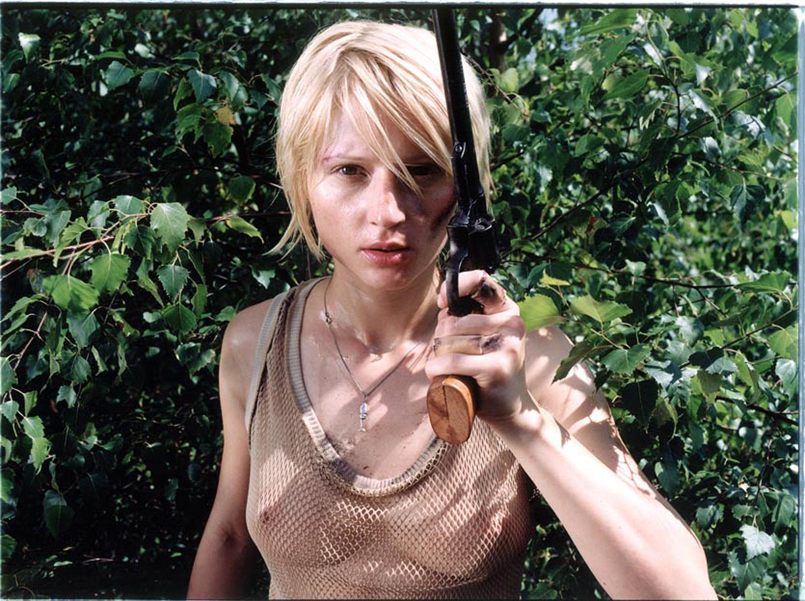 A woman with a short, blonde haircut and a tank top, holding a gun in a forest-like setting.