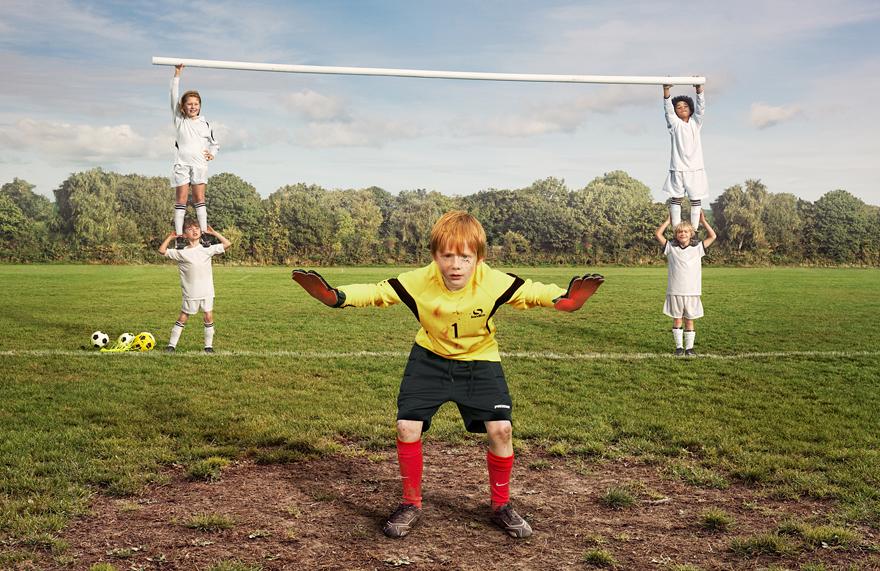 A young boy in a yellow football jersey and black shorts is standing in a field, surrounded by a group of children holding a large white goalpost.
