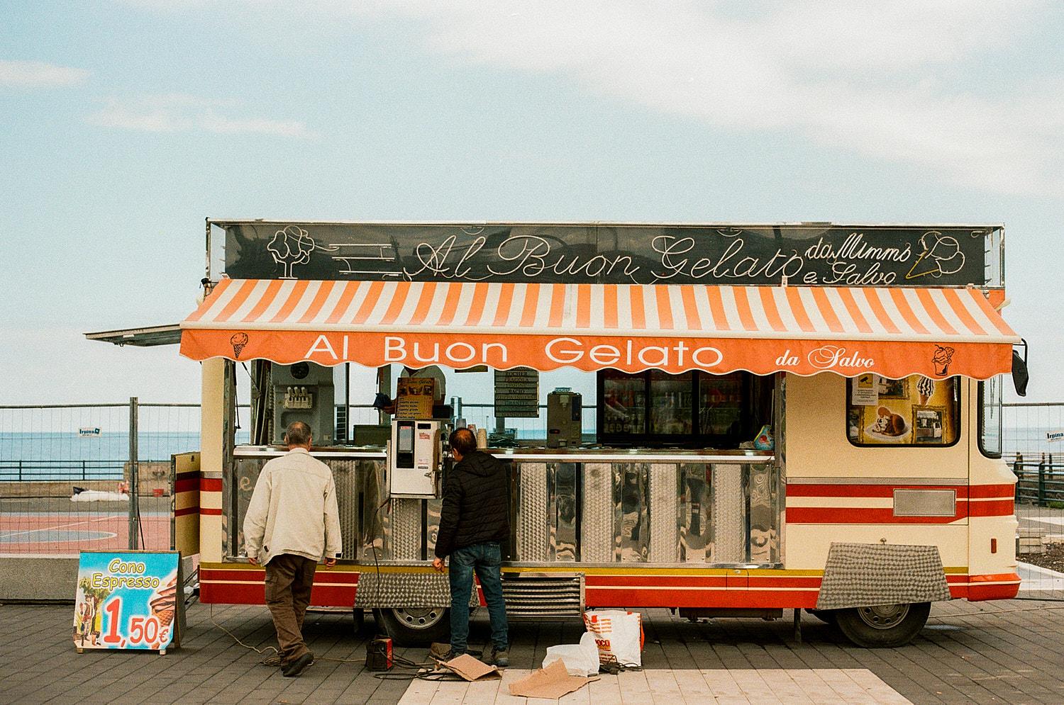 A food truck, likely a gelato truck, is parked on a beach or waterfront, with a sign in Italian and a menu board in the window.