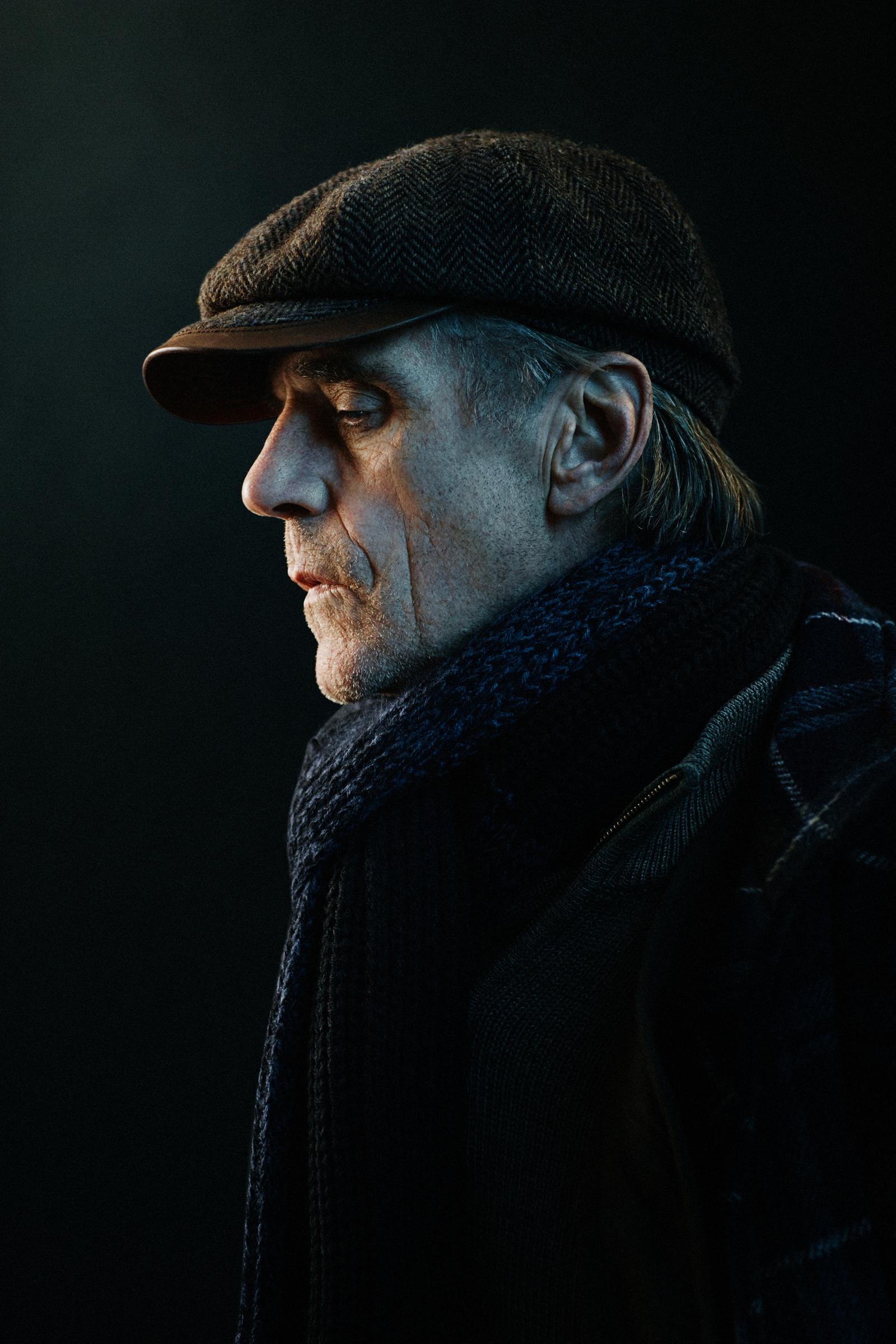 A man in his 60s with grey hair, wearing a flat cap, a dark coat, and a scarf, looking to the side with a serious expression.