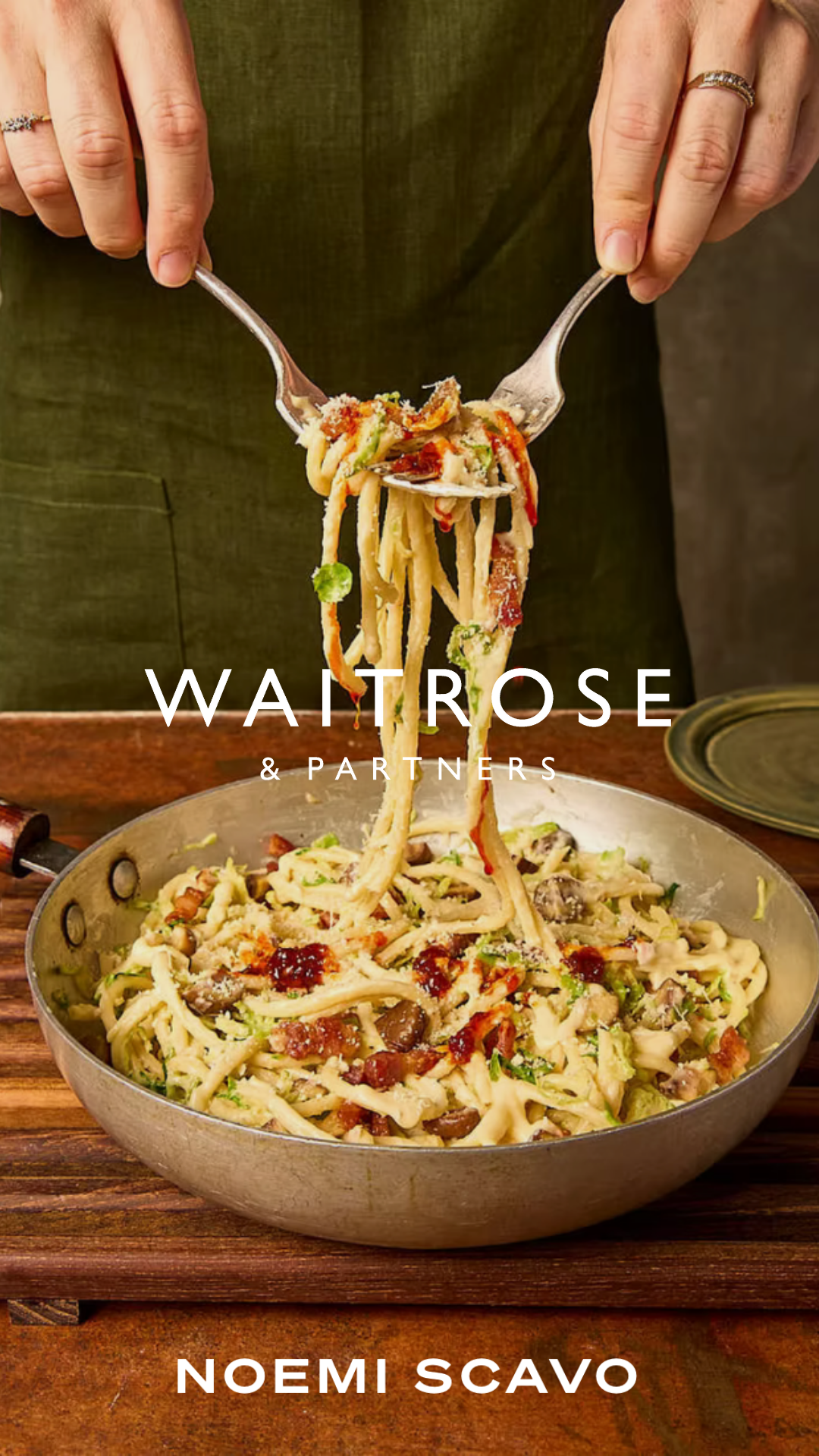 The image shows a person in a green apron using two forks to lift a large portion of creamy pasta from a silver skillet. The pasta is mixed with pancetta and greens, and the shot is framed as a promotional graphic for Waitrose by Noemi Scavo.