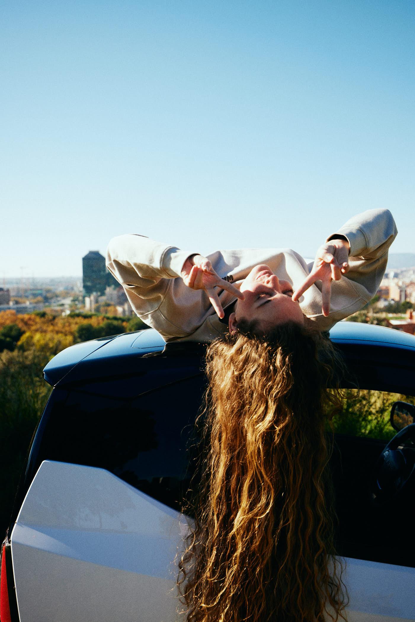 A woman with long, curly hair is leaning out of the open sunroof of a white car, making a peace sign with her hands. She is upside down, looking directly at the camera with a.