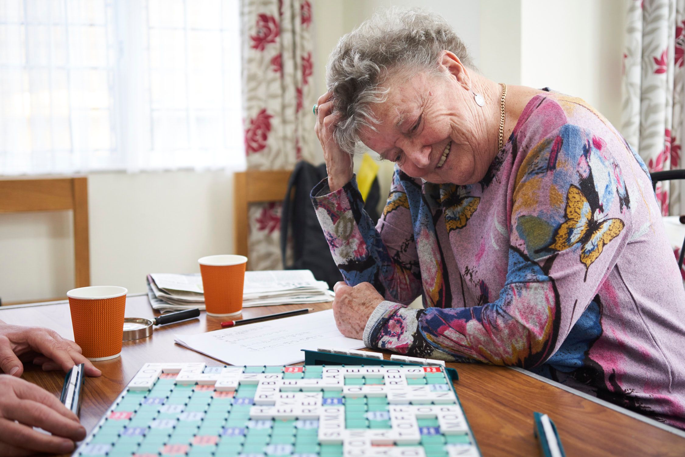 An elderly woman with grey hair, wearing a colourful floral blouse, sitting at a table with a Scrabble board, looking frustrated and holding her head in her hand.