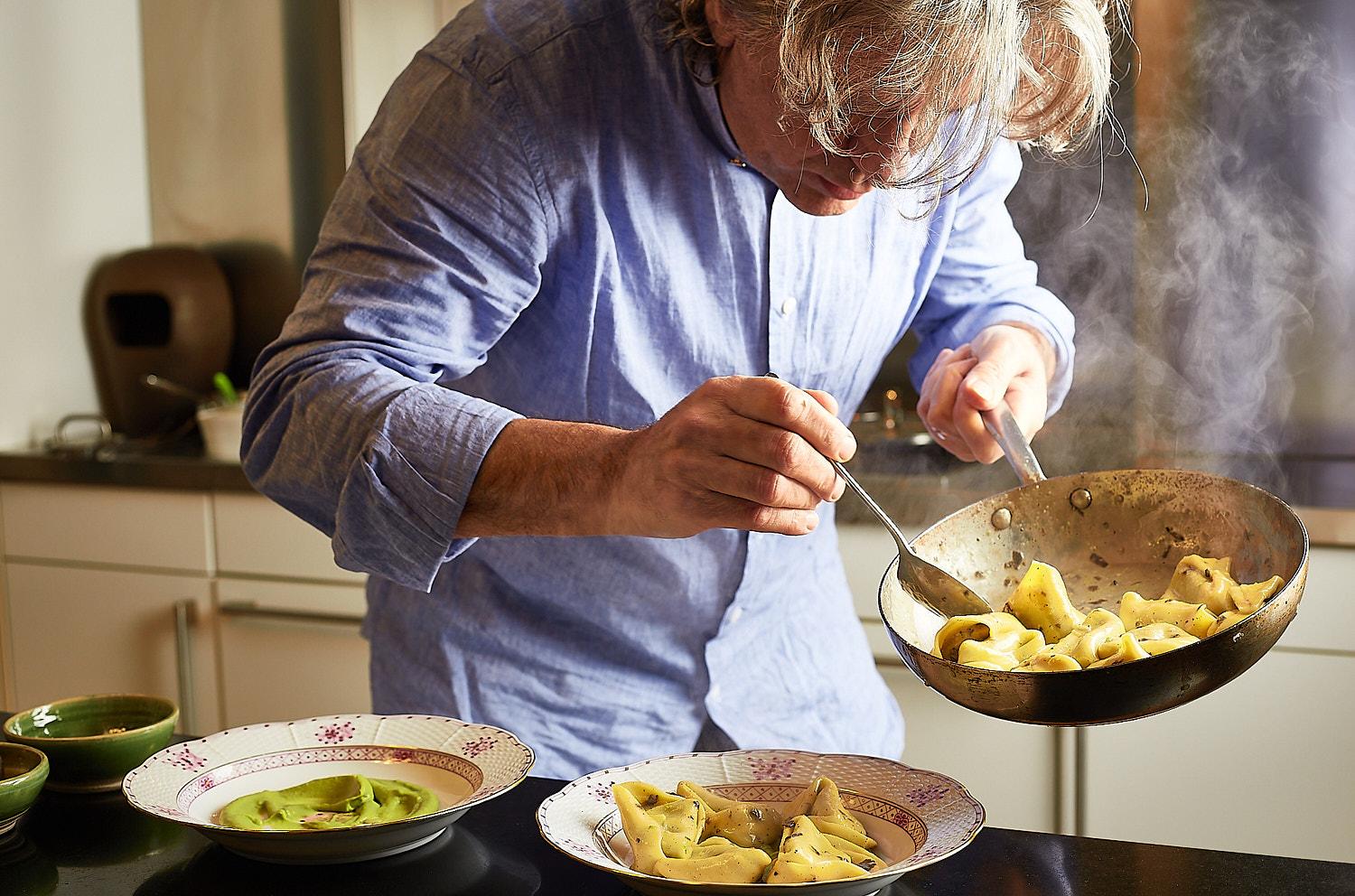 A chef, likely Noemi Scavo, is cooking in a kitchen, stirring a large pan of pasta with a wooden spoon.