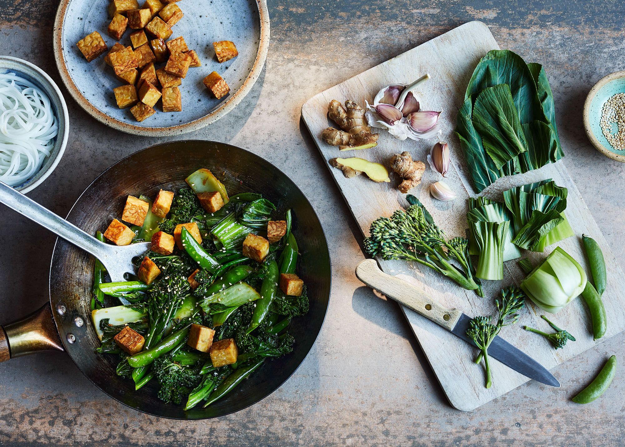 A wok with a stir-fry of vegetables and tofu, with a few ingredients on a cutting board nearby.
