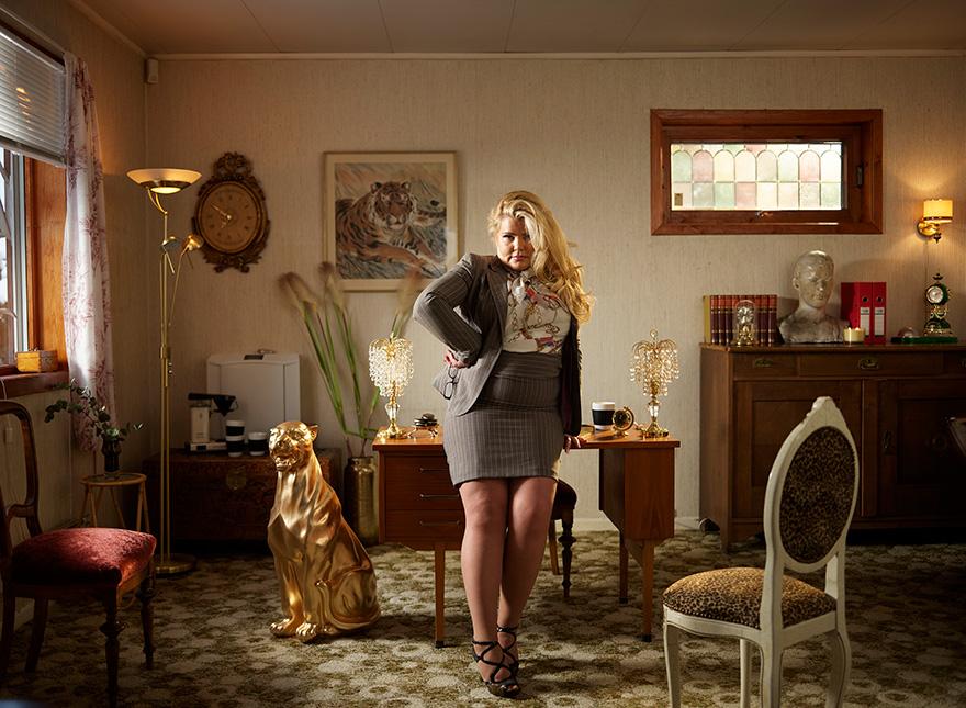 A woman in a business suit, standing in a dimly lit, retro-style office with a tiger painting on the wall, a gold cat statue, and a tiger-shaped clock.