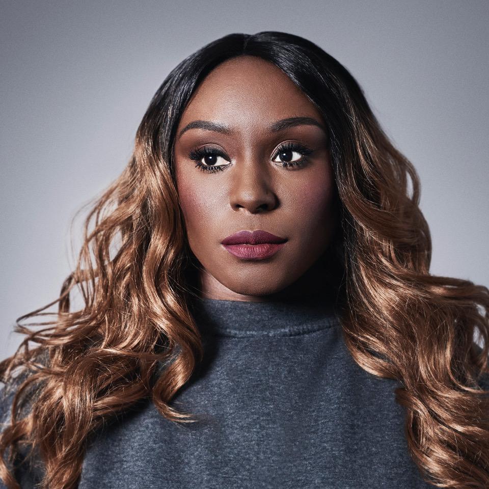A woman with long, curly brown hair and a grey, high-necked, long-sleeved top, looking directly at the camera with a serious expression.