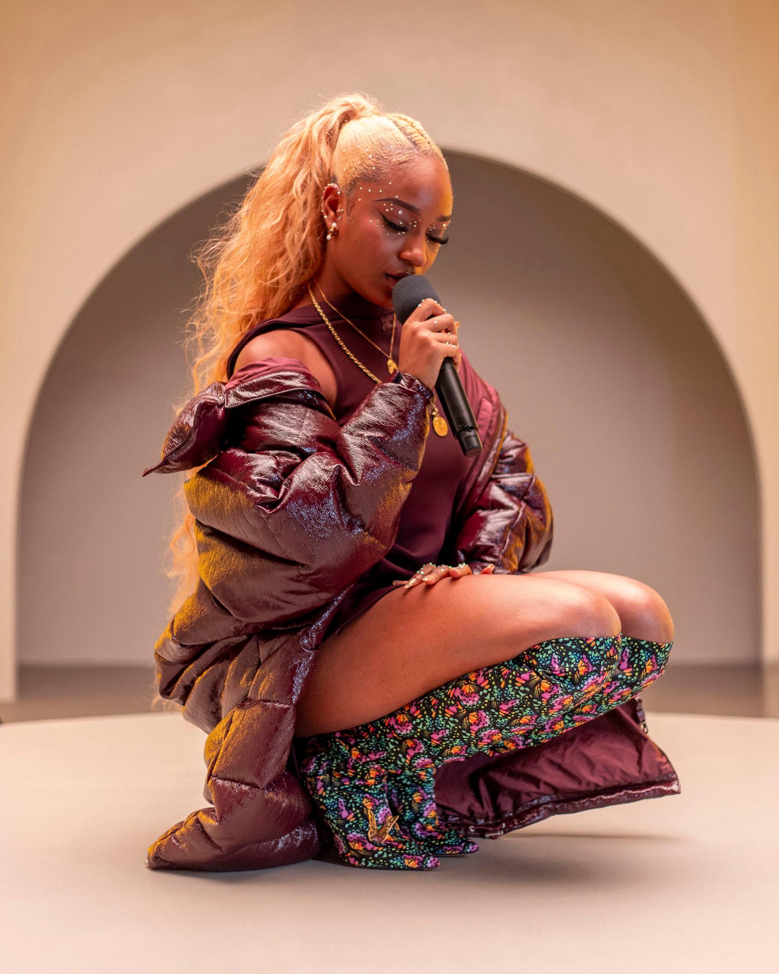A woman with long blonde hair and a maroon puffy jacket, holding a microphone and sitting on the floor.