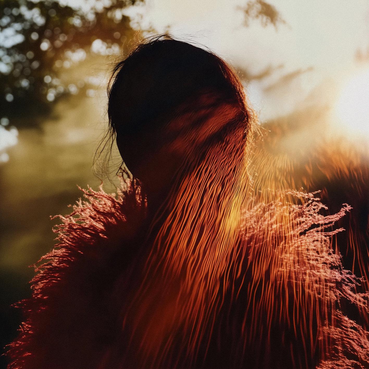 A woman with long, flowing hair and a subtle, ethereal glow, standing in a lush, natural environment with trees and foliage in the background.