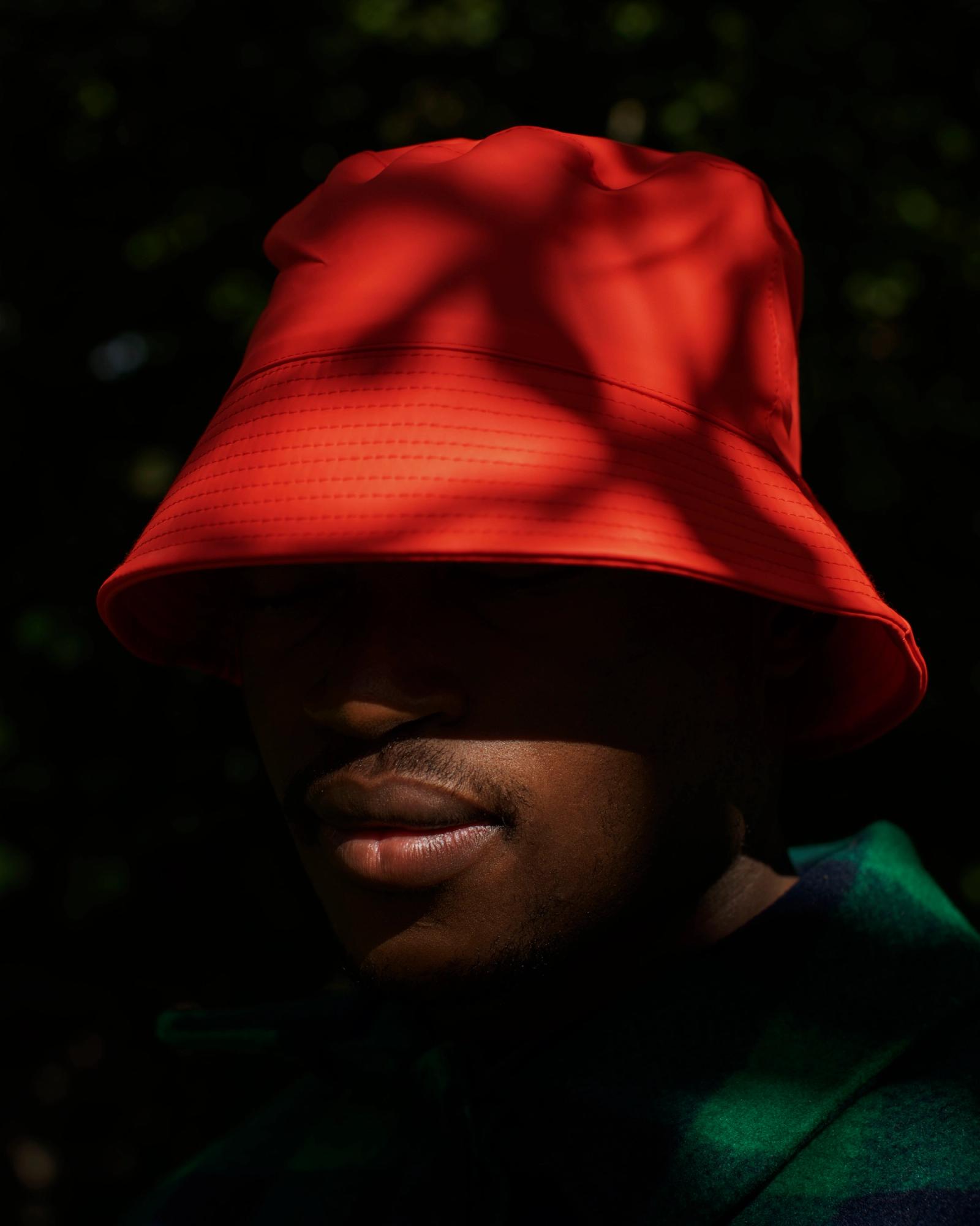 A person wearing a red bucket hat is standing in front of a blurred background, with the hat's brim casting a shadow on their face.