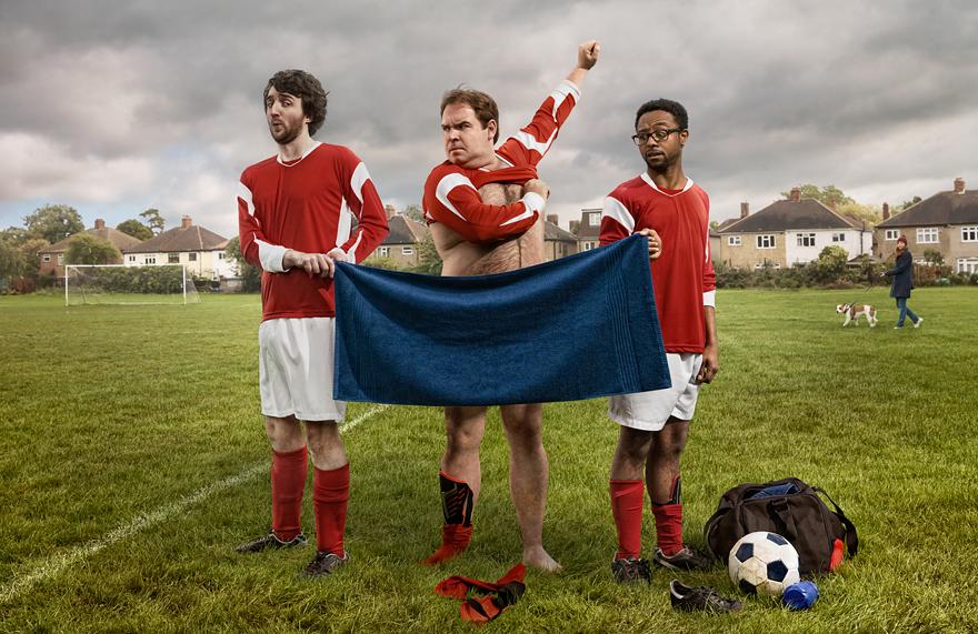 A group of men in football jerseys and shorts, one of them holding a blue towel, standing on a grassy field with a goal in the background.