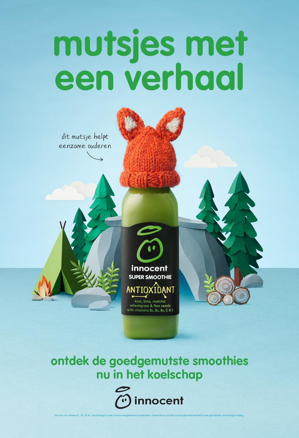 A green smoothie bottle with a knitted orange fox hat on top, surrounded by a campfire, a tent, and a few trees in a forest-like setting.