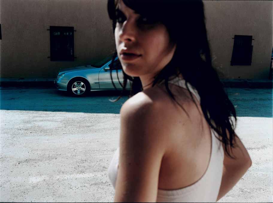 A woman in a white tank top and jeans, standing in a parking lot with a silver car in the background.
