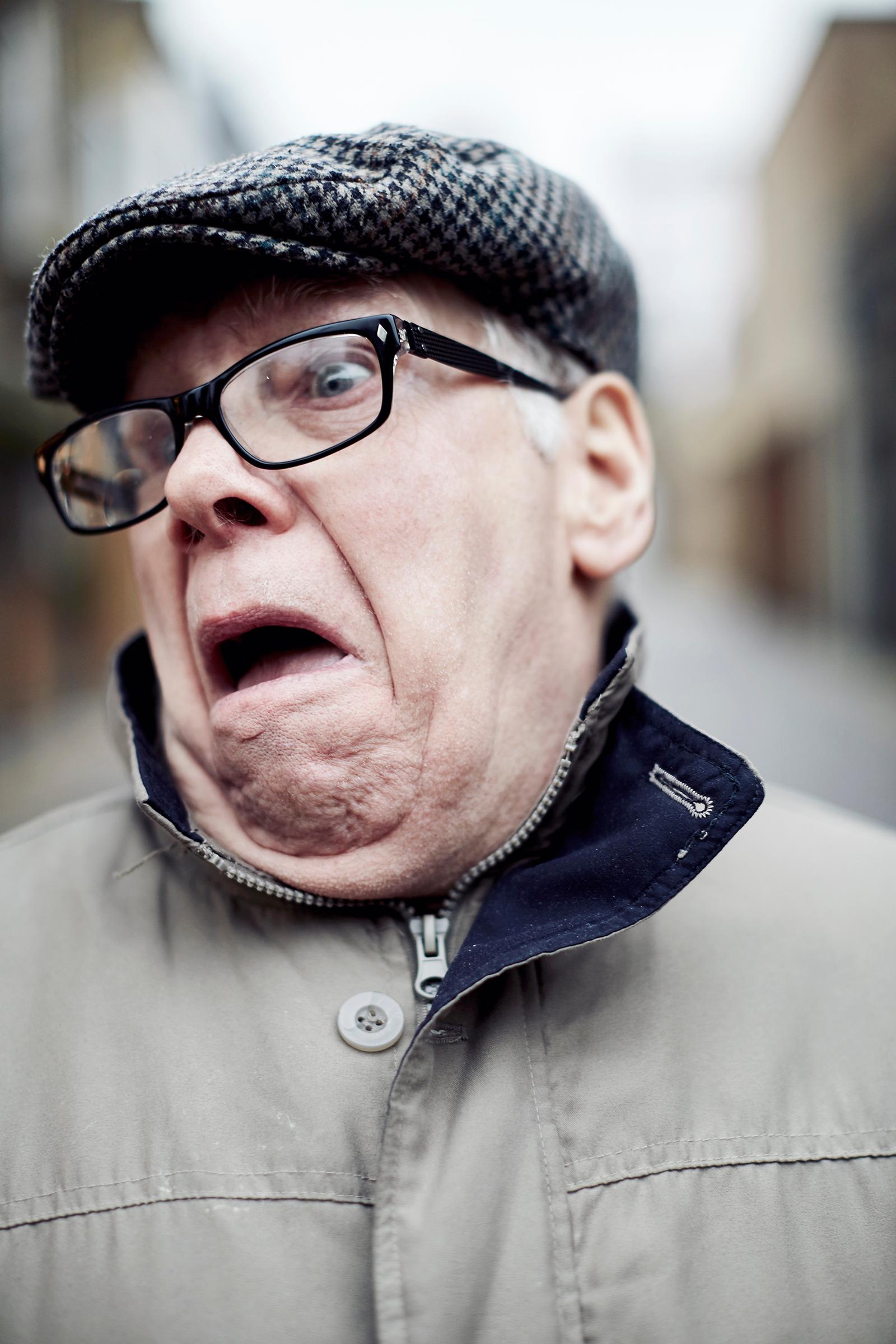 An elderly man with a shocked expression, wearing a flat cap and a thick, grey, woolen coat, with a blurred background.