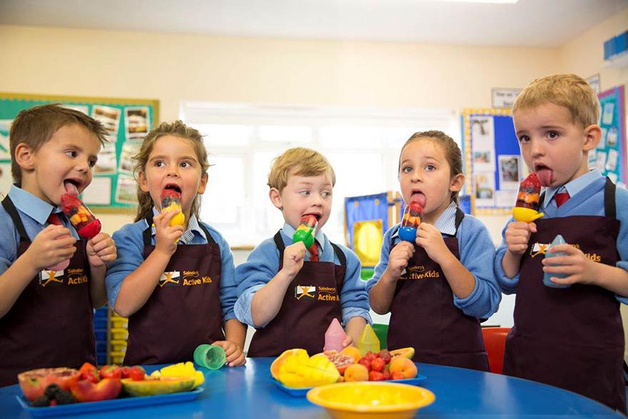 A group of children in Sainsbury's Active Kids uniforms are eating fruit kebabs in a classroom.
