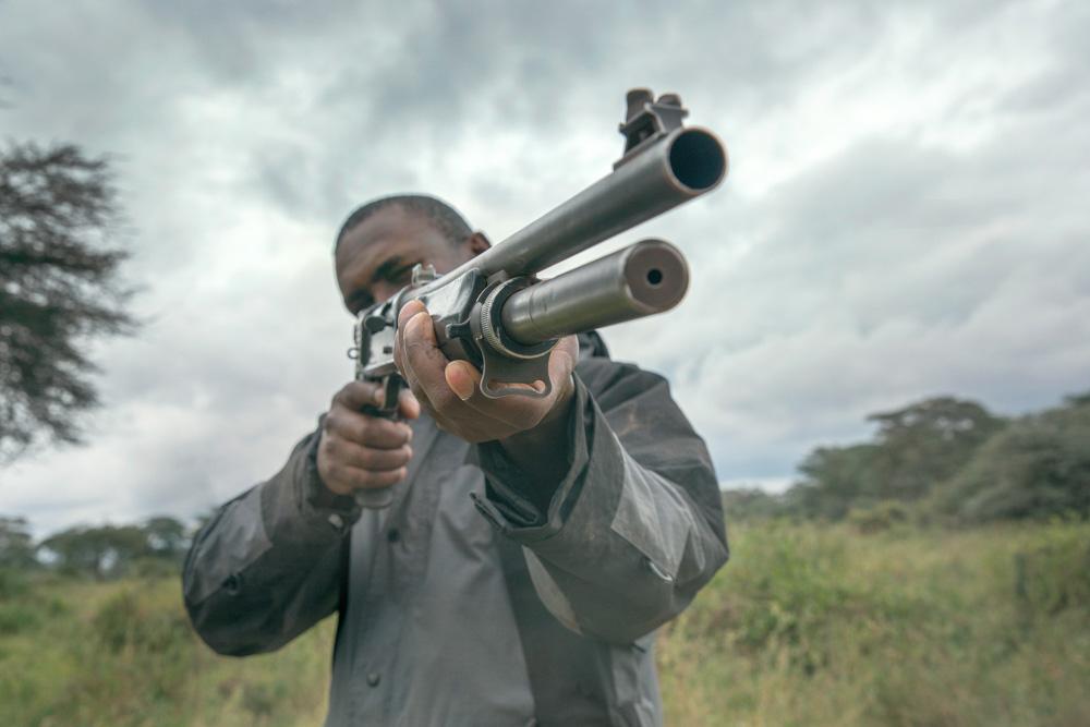 A man in a black jacket and hat holding a rifle, aiming at something in the distance, in a grassy savannah landscape.