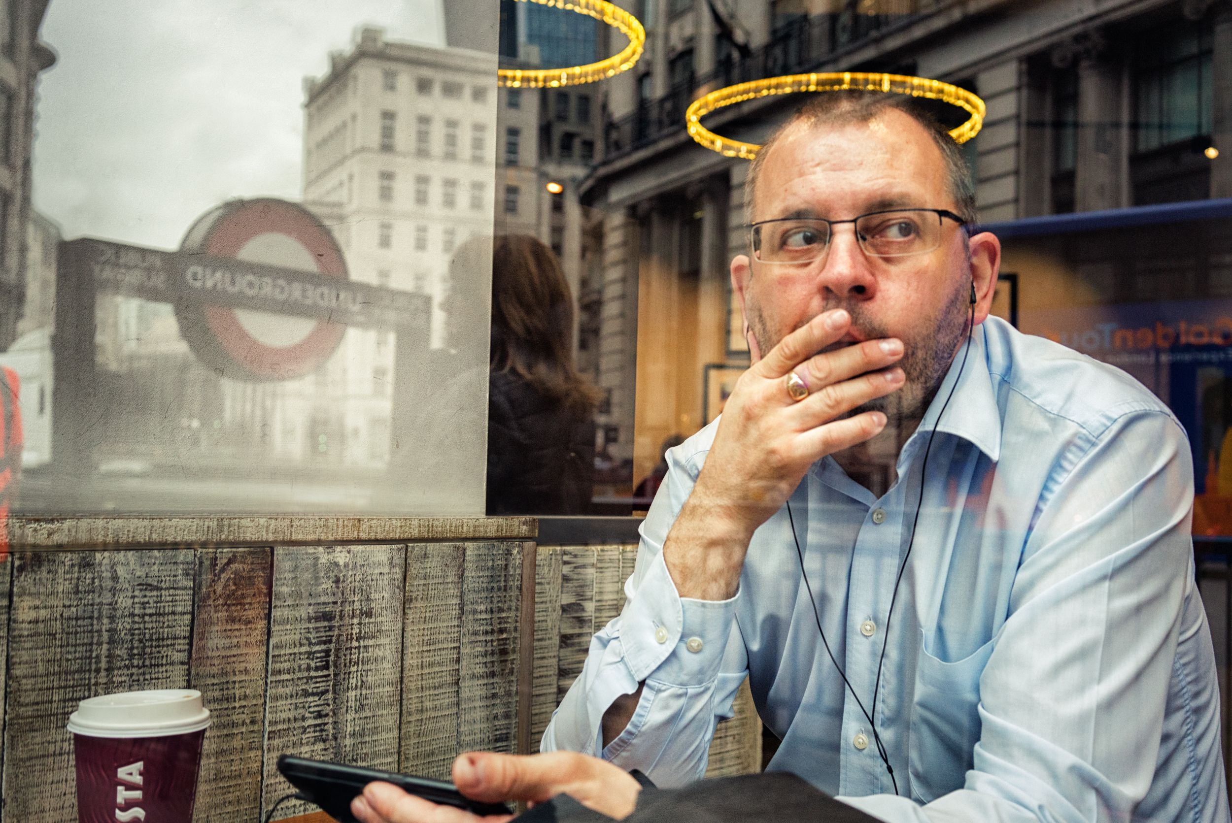 A man sits in a coffee shop, looking out the window at a London Underground sign.