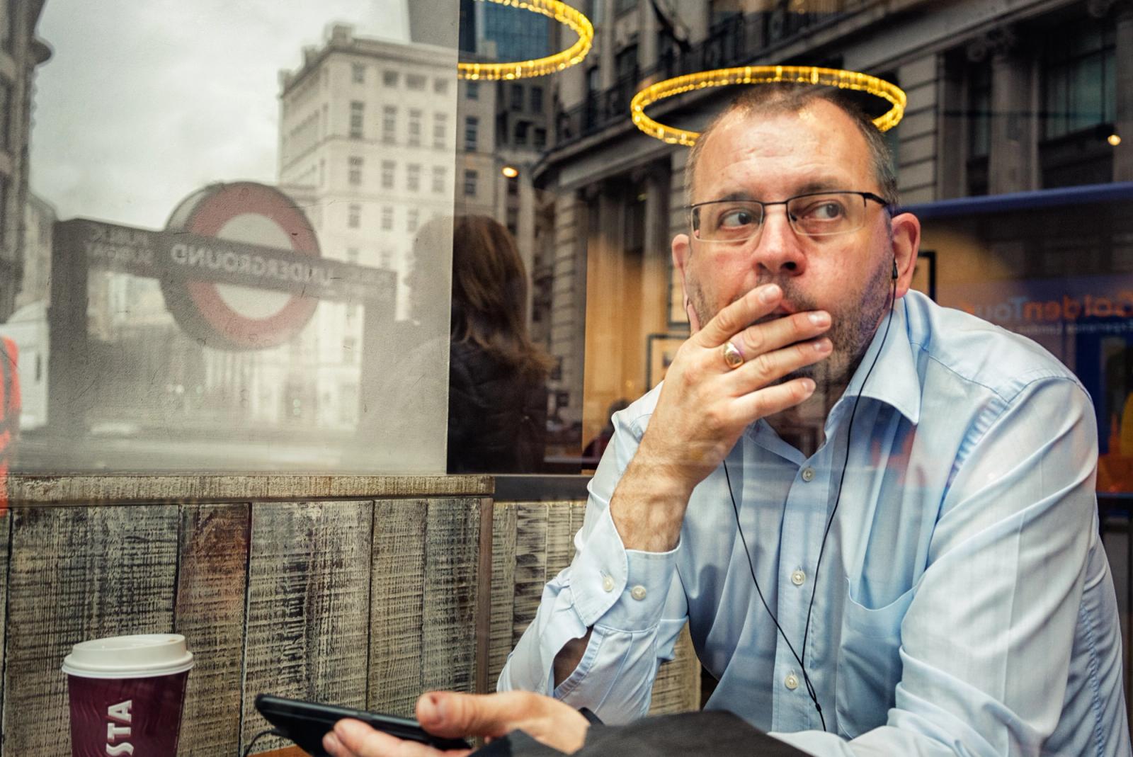A man sits in a coffee shop, looking out the window at a London Underground sign.