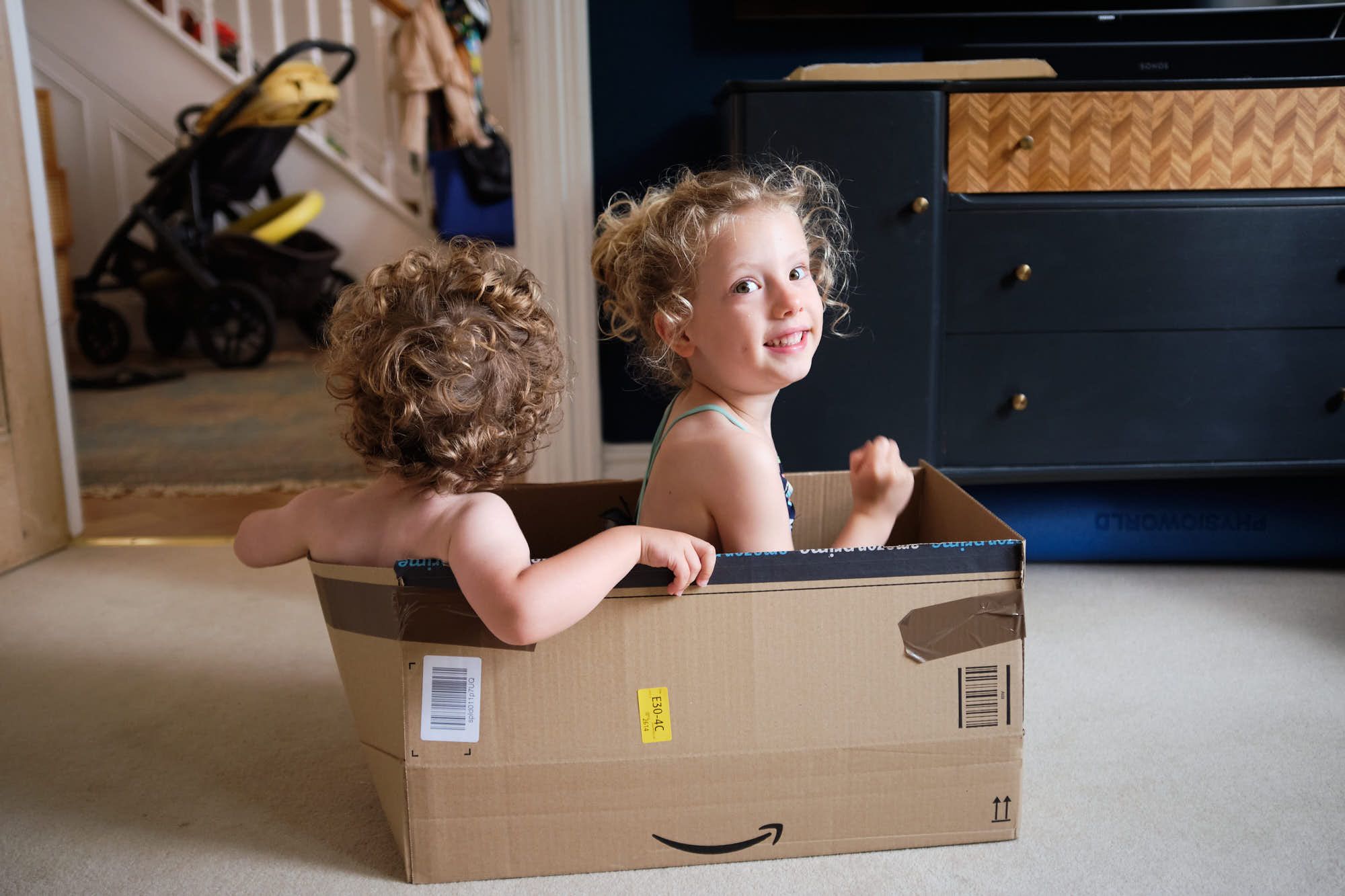 Two young children, likely twins, playfully sit in a large cardboard box, their curly hair and wide eyes capturing a sense of joy and playfulness. The box, with its Amazon logo.