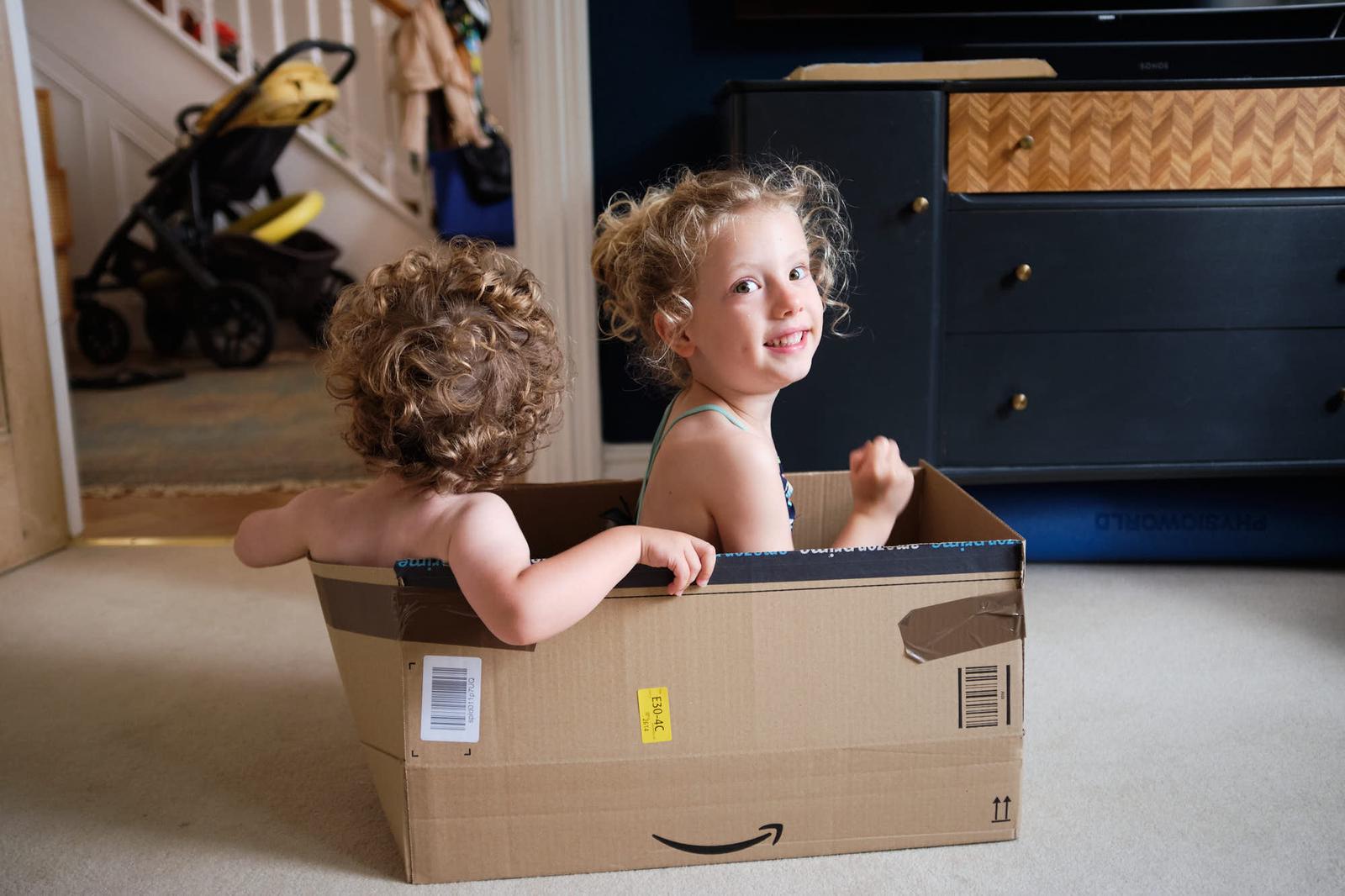 Two young children, likely twins, playfully sit in a large cardboard box, their curly hair and wide eyes capturing a sense of joy and playfulness. The box, with its Amazon logo.