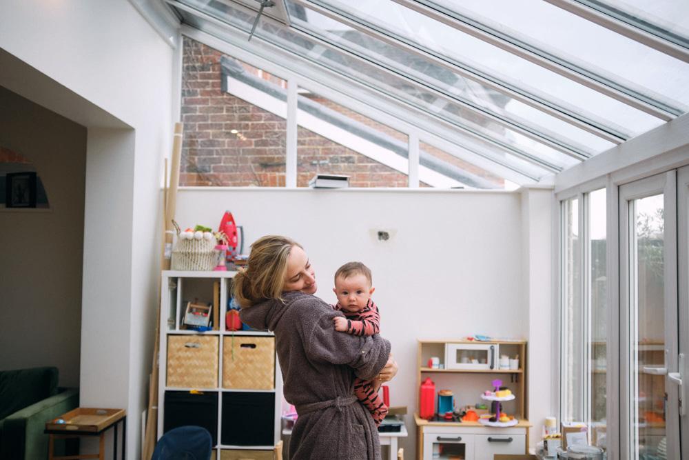 A woman in a bathrobe holds a baby in a playroom. The woman is looking at the camera with a neutral expression, while the baby is looking away with a serious expression.