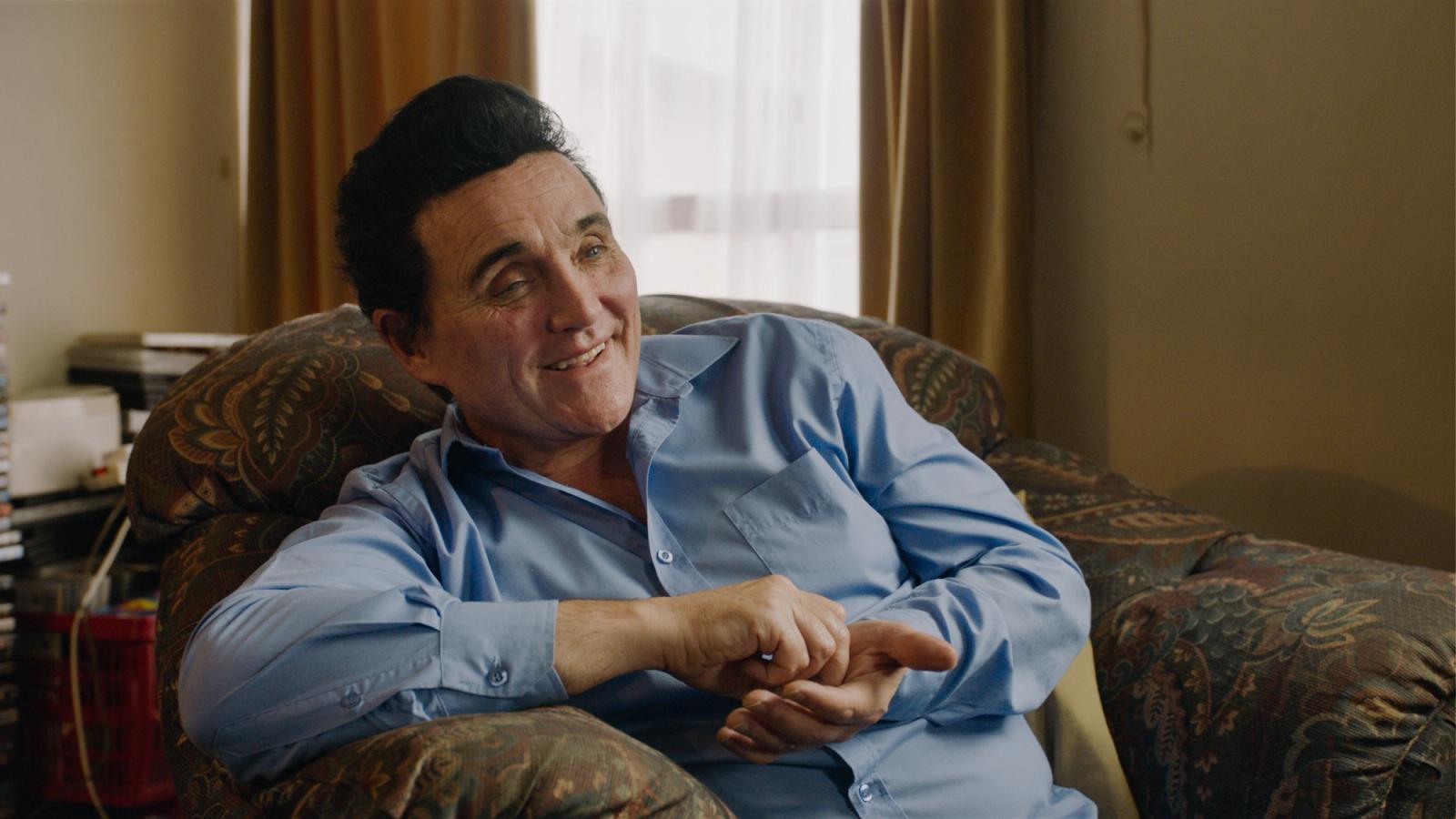Callum O'Keefe Still from JUAN film, about elvis impersonator blue shirt smiling talking on sofa