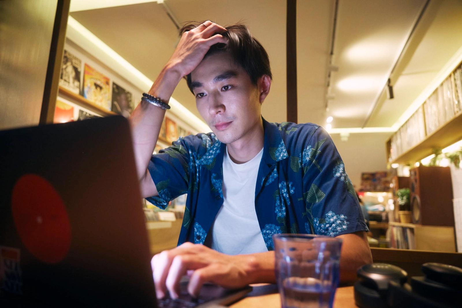 A young Asian man with his hand on his forehead, sitting in front of a laptop in a dimly lit record store. He appears to be stressed or frustrated.