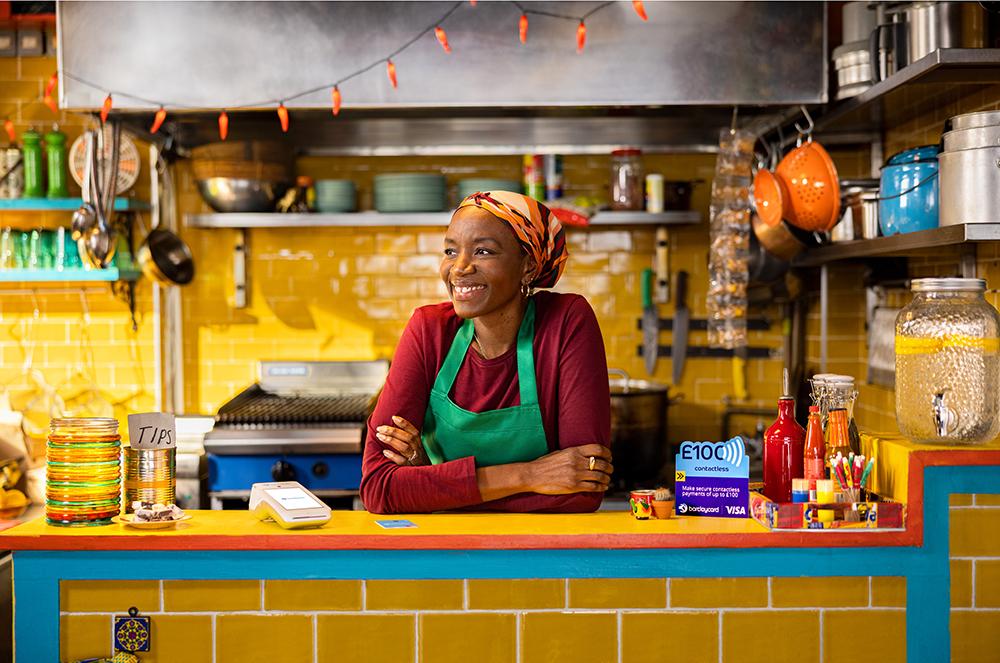 A woman in a red shirt and green apron is standing behind a counter in a kitchen, surrounded by various food and cooking equipment.