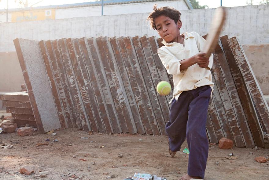 A young boy is playing cricket with a tennis ball in a rough, outdoor setting.