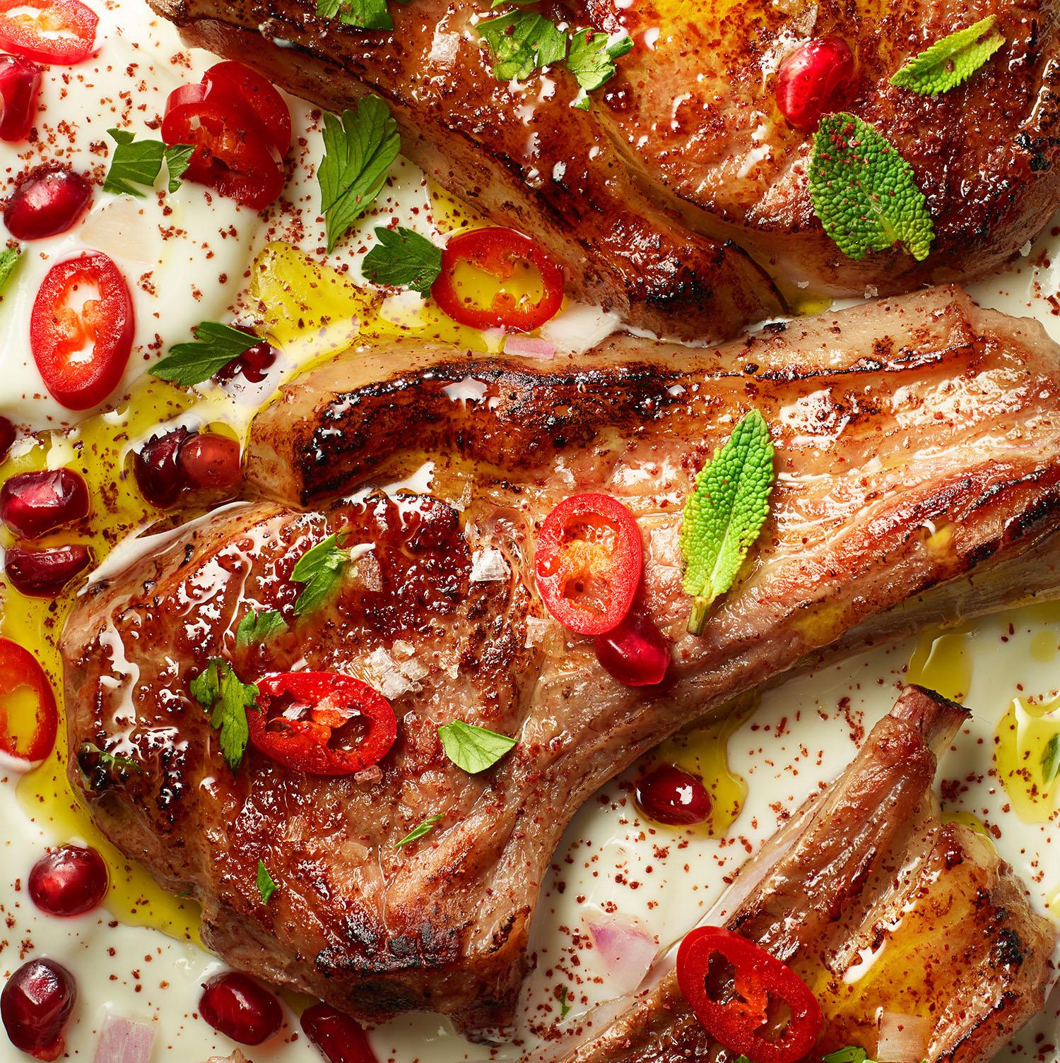 A close-up of lamb chops on a plate with pomegranate, red chili, and mint.