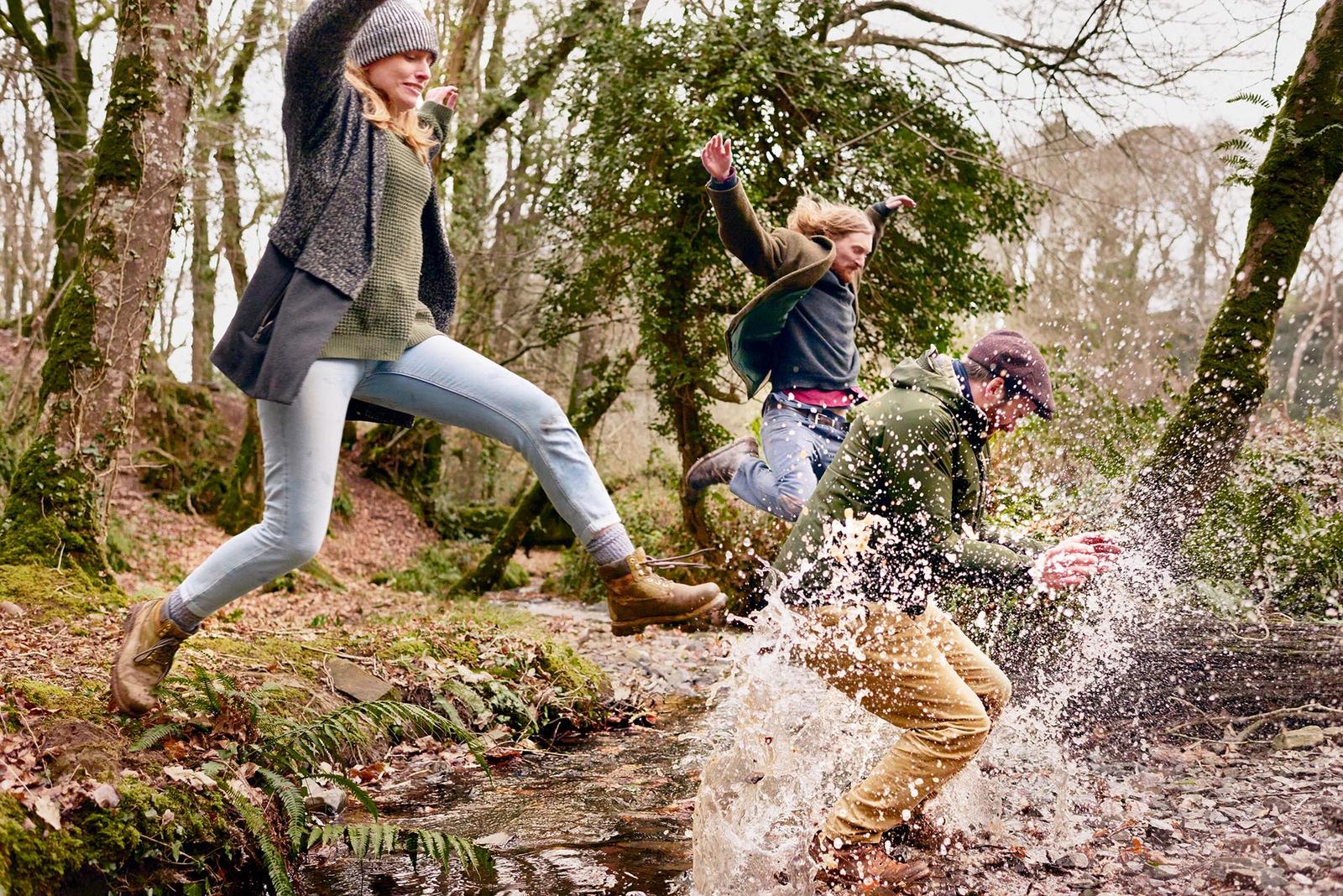 Three people, two women and one man, jumping over a small stream in a forest, wearing hiking boots, jeans, and warm clothing, including a hat and scarf.