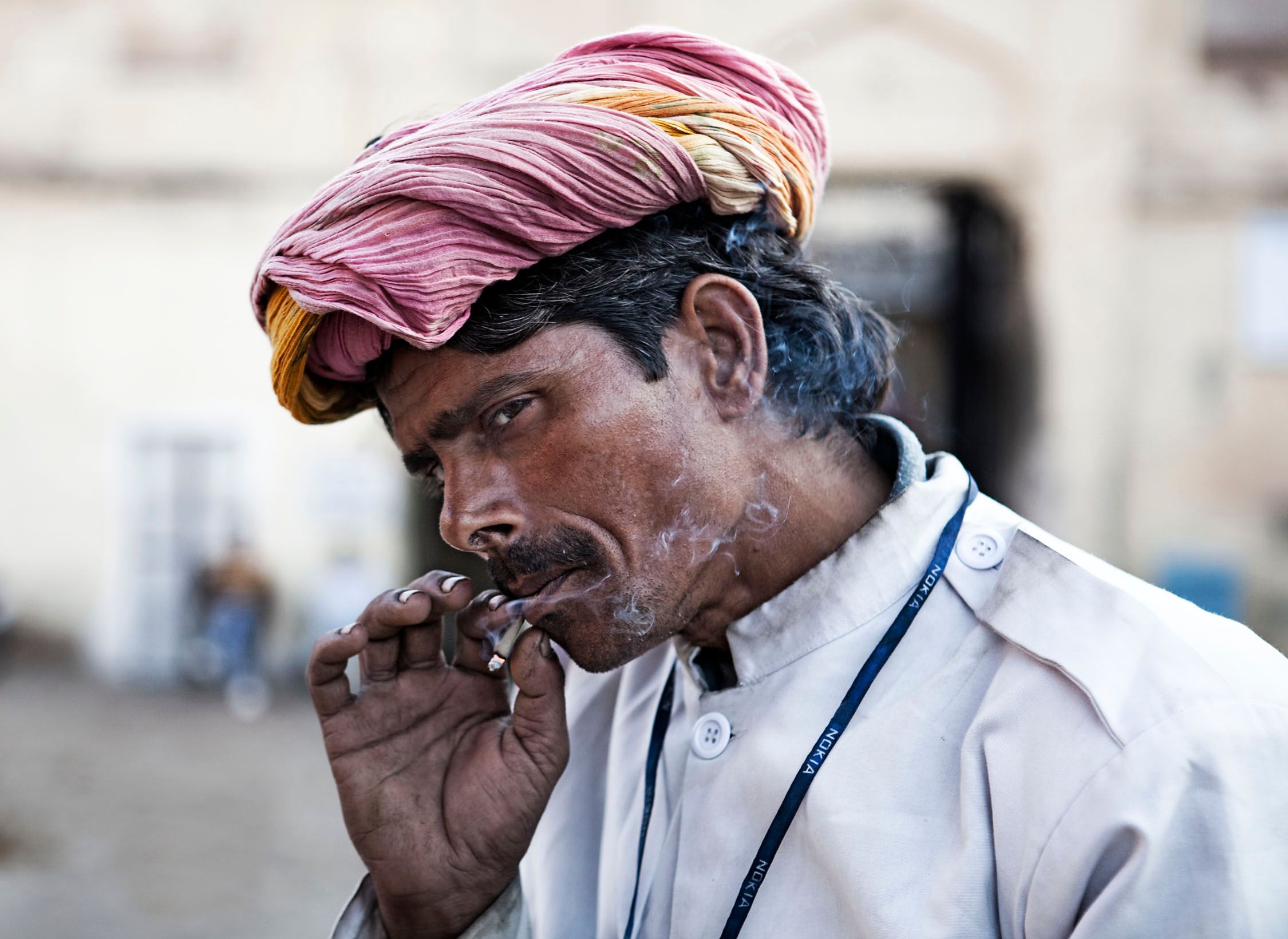 A man in a white shirt and pink turban smokes a cigarette, with a serious expression on his face. He is wearing a blue lanyard with white text, possibly indicating his role or.