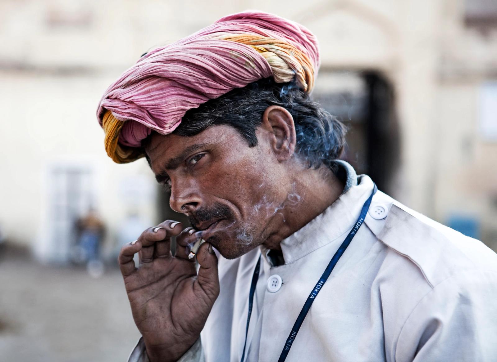 A man in a white shirt and pink turban smokes a cigarette, with a serious expression on his face. He is wearing a blue lanyard with white text, possibly indicating his role or.