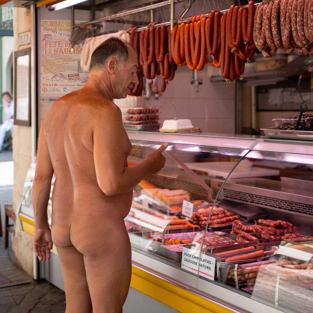 A man, naked, standing in front of a meat counter, examining various types of sausages and meats. AI by Alys Thomas