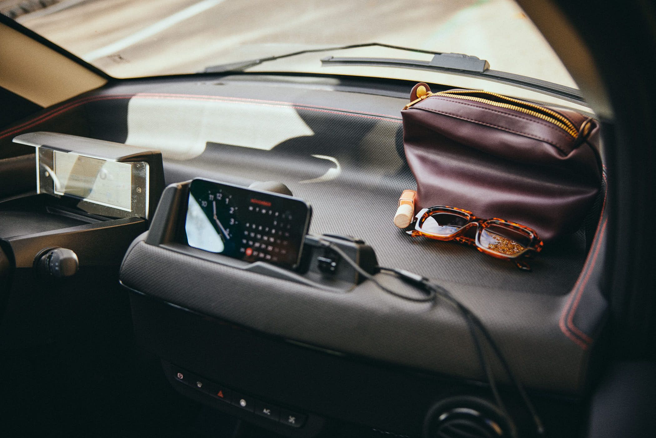 A dashboard of a car, with various items on it. A pair of glasses, a small pouch, and a phone are arranged on the dashboard, suggesting a driver's readiness to drive.