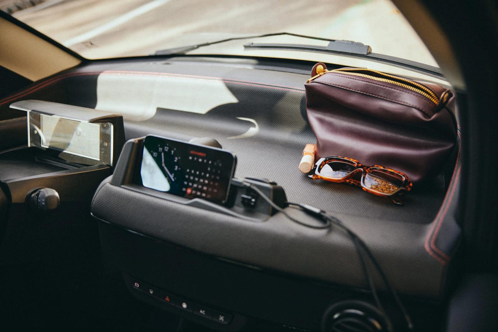 A dashboard of a car, with various items on it. A pair of glasses, a small pouch, and a phone are arranged on the dashboard, suggesting a driver's readiness to drive.