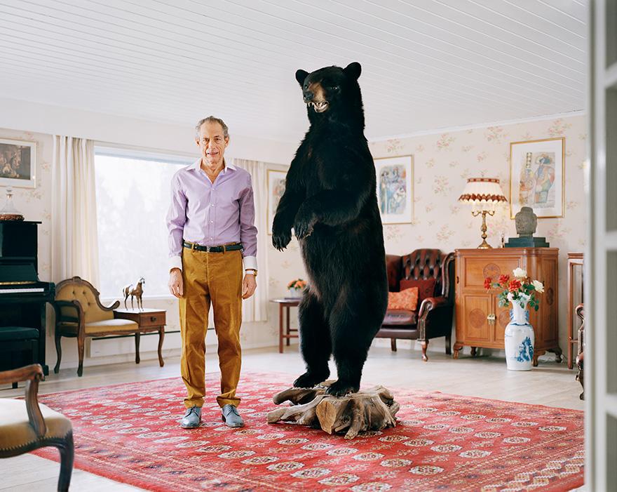 A man in a purple shirt and brown trousers standing next to a large black bear in a living room.
