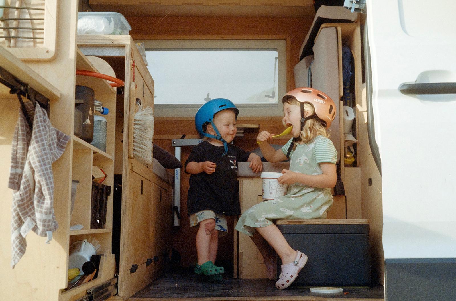 Two children in a van, both wearing helmets and eating ice cream.