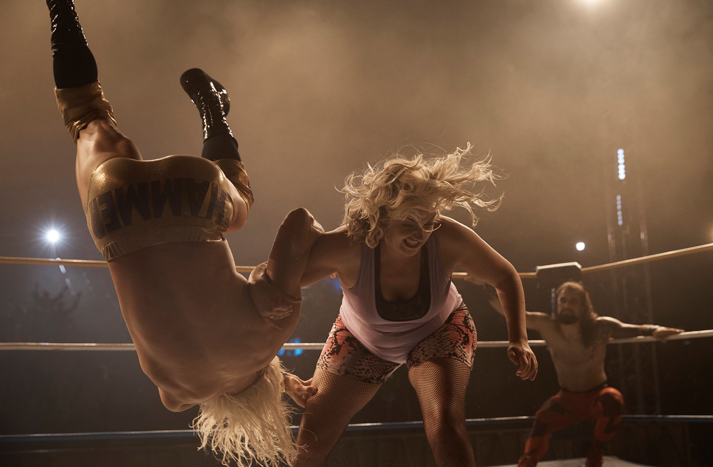 A female wrestler in a pink tank top and leopard-print shorts is being thrown by a male wrestler in a gold and black wrestling costume, with a crowd in the background.